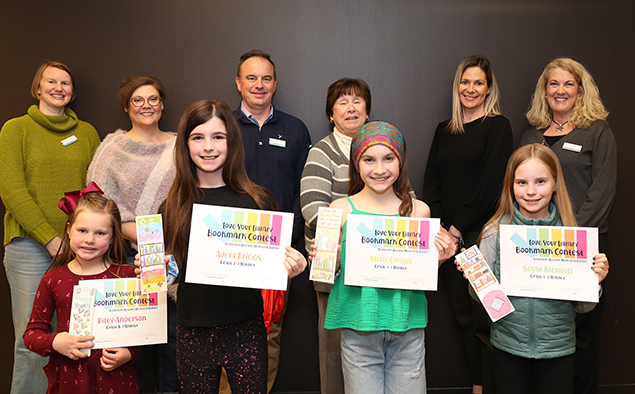 Four students hold their certificates and winning bookmarks while standing in front of library board members