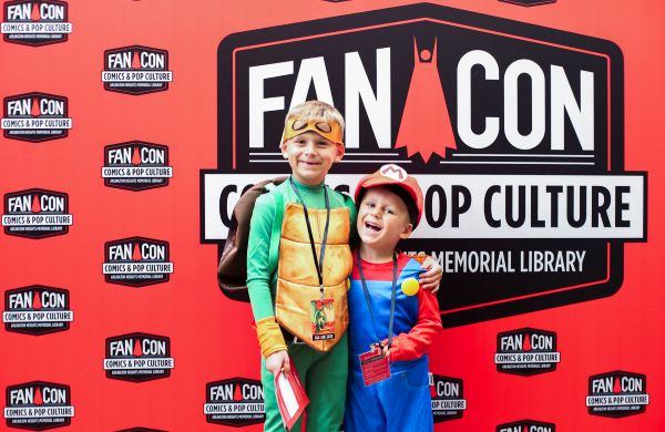 Two boys in cosplay. One Teenage Mutant Ninja Turtles Cosplay and the other in a Mario from Super Mario Brothers Cosplay. They stand in front of an AHML FanCon Step-and-repeat.